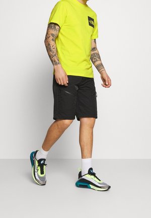 Mand, der går, iført en knaldgul North Face T-shirt, sorte shorts med lynlåslommer og sneakers med neonfarvede detaljer samt hvide sokker.