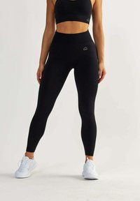 Schwarze Leggings aus dehnbarem Stoff, hochgeschnittenes Design und ein kleines Herz-Logo. Kombiniert mit einem schwarzen Sport-BH und weißen Sneakers.