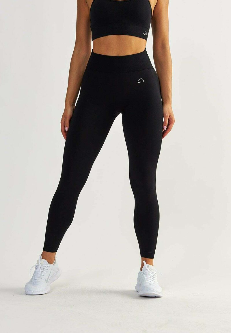 Schwarze Leggings aus dehnbarem Stoff, hochgeschnittenes Design und ein kleines Herz-Logo. Kombiniert mit einem schwarzen Sport-BH und weißen Sneakers.