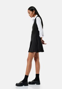 Zwarte geplooide jurk met een aansluitende bodice, gelaagd over een witte blouse. Heeft een zilveren accent bij de taille. Zwarte schoenen met dikke zolen.
