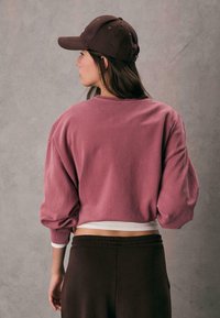 Roza oversized dukserica s dugim rukavima, rebrastim manžetama i bijelim rubom. Uparena s tamnobraon hlačama s elastičnim pojasom i smešnim kapom.
