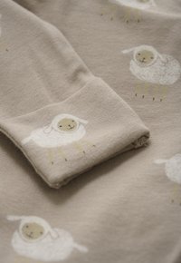 Zacht beige stof met een opgevouwen mouw, versierd met een herhaald patroon van kleine, lachende witte lammetjes met lichtgele poten.