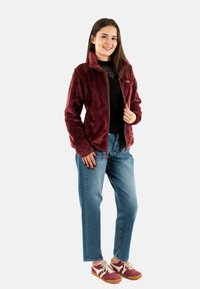 Giacca in fleece bordeaux con zip frontale, tasche laterali e texture decorata; abbinata a jeans blu e scarpe sportive bordeaux.