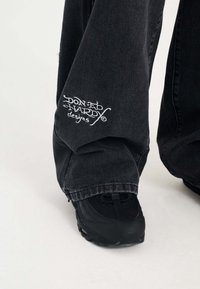 Musta farkut, joissa on vasemmassa lahkeessa brodeeratut "Don Ed Hardy designs", yhdistettynä mustiin urheilukenkiin, korostaen rentoa istuvuutta.