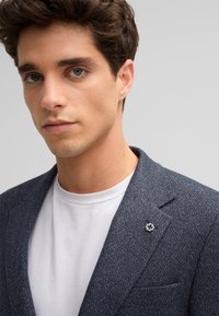 Marineblauer strukturierter Blazer mit Revers, mit einer dekorativen Brosche und einem weißen Rundhals-T-Shirt darunter. Sauberes, modernes Design.
