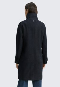 Donker wollen jas met een hoge kraag, voorzien van een visgraatpatroon, knoopmanchetten en een enkele haaksluiting op de rug.