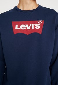 Mörkblå sweatshirt i bomull med rund halsringning, utrustad med en stor röd Levi's-logotyp på bröstet i vita bokstäver.