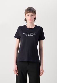 Juodas medvilninis marškinėliai su apvalia iškirpte ir trumpomis rankovėmis. Balti užrašai skaito "Marc O’Polo EST. IN STOCKHOLM" priekyje.