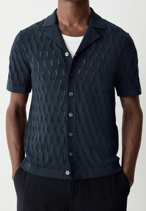 Homme portant un pull en maille bleu foncé à manches courtes avec un motif texturé et des boutons sur un sous-vêtement blanc, mains dans les poches.