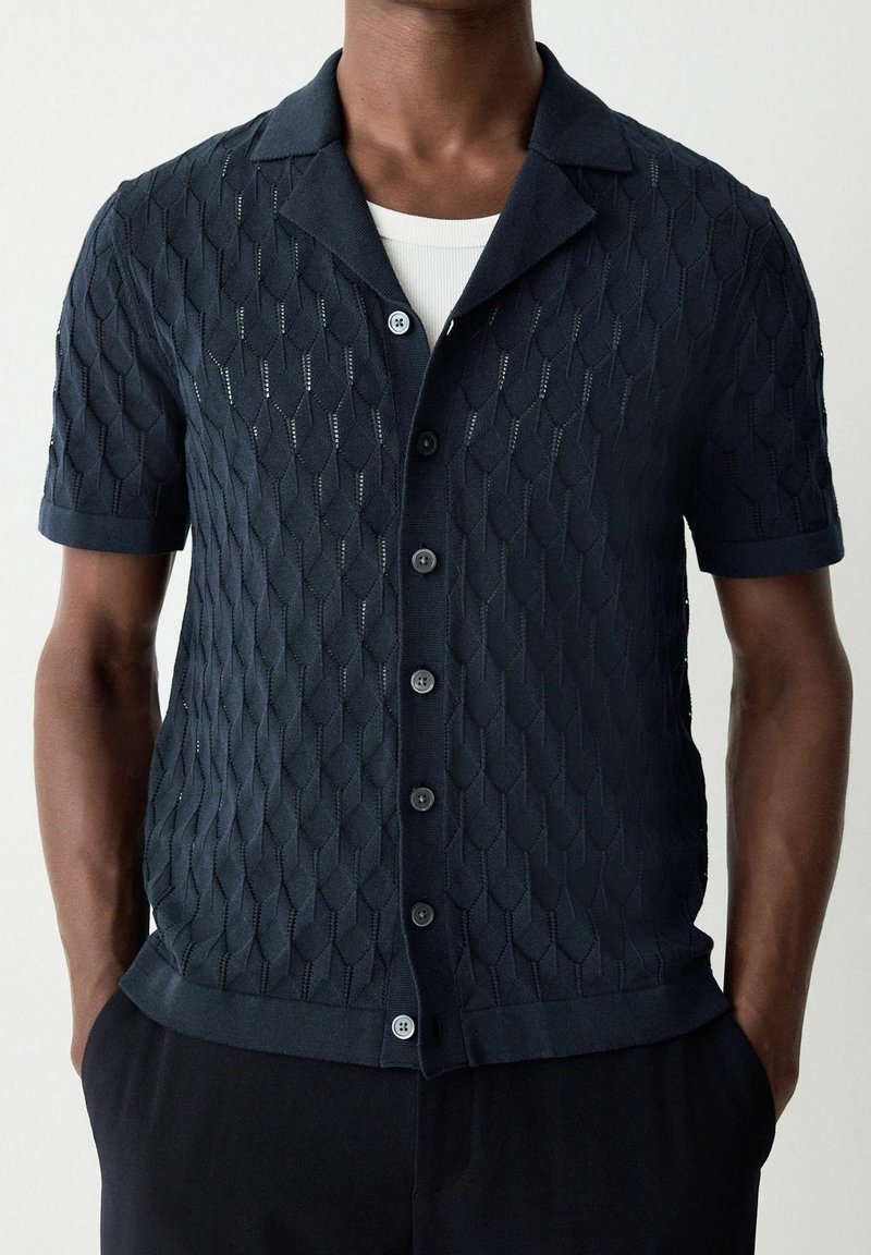 Homme portant un pull en maille bleu foncé à manches courtes avec un motif texturé et des boutons sur un sous-vêtement blanc, mains dans les poches.