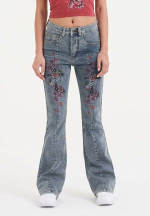 Jeans flare in denim blu sbiadito con ricami floreali e a croce sul davanti, abbinati a un top rosso corto e sneakers bianche.