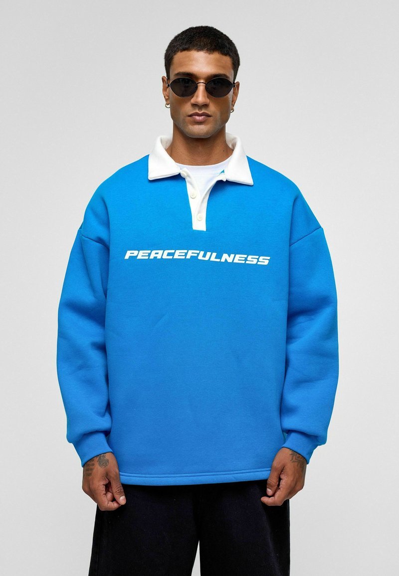 Blauwe sweatshirt met witte kraag en grote tekst "VREDE". Gemaakt van een zachte stof; heeft lange mouwen en een losse pasvorm.