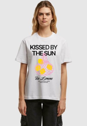 Weißes Baumwoll-T-Shirt mit schwarzem Text "VON DER SONNE GEKÜSST" und Grafik von Zitronen in einer pinken Netztasche. Kurze Ärmel, lockere Passform.