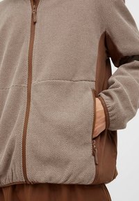 Lett brun jakke med glidelås som har et strukturert mesh-materiale, solide brune detaljer og to sidelommer.
