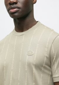 Camiseta verde oliva claro con estampado tonal "EA7 Emporio Armani" en toda la prenda y un pequeño logotipo metálico en el área del pecho izquierdo.