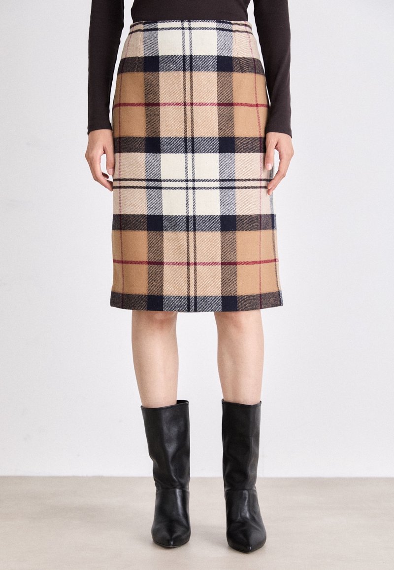 Femme portant une jupe à carreaux beige, noire, blanche et rouge arrivant au genou, avec des bottes en cuir noires mi-mollet, debout devant un fond uni.