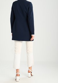 Cappotto tailored blu navy con silhouette aderente, maniche lunghe e tessuto liscio, abbinato a jeans bianchi a vita alta e svasati e tacchi bianchi.
