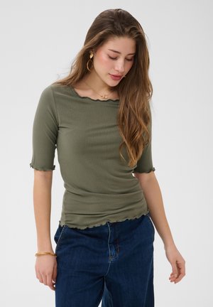 Ung kvinde med langt hår bærer en olivengrøn ribbet top og mørkeblå jeans, kigger ned med hænderne tæt på lommerne mod en enkel baggrund.