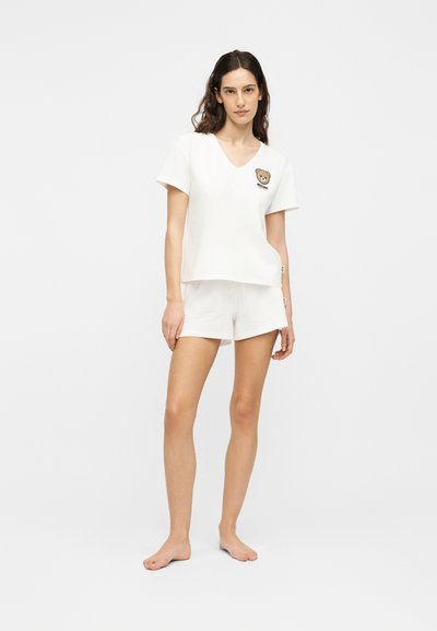 Femme debout pieds nus portant un haut blanc à manches courtes et un short, le haut présente un petit motif d'ours en peluche près de l'épaule.