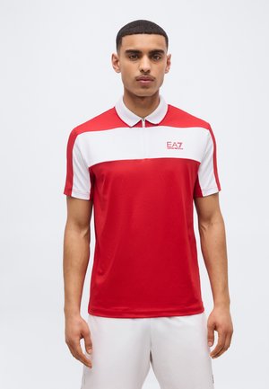 Mann steht, trägt ein rot-weißes EA7 Emporio Armani Polohemd mit kurzen Ärmeln und weiße Shorts vor einem einfarbigen Hintergrund.