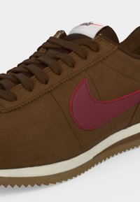 Καφέ αθλητικό παπούτσι Nike με σκουροκαφέ κορδόνια, κόκκινο λογότυπο Swoosh, λευκή μεσαία σόλα και υφασμένη καφέ εξωτερική σόλα.