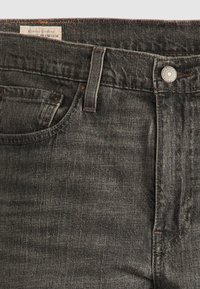 Gros plan d'un jean en denim noir montrant la poche avant, la boucle de ceinture, le bouton et une étiquette de marque partiellement visible à l'intérieur de la ceinture.