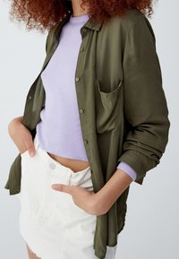 Chemise à manches longues vert olive avec boutons et poche poitrine, superposée à un pull violet clair, associée à une mini-jupe blanche.
