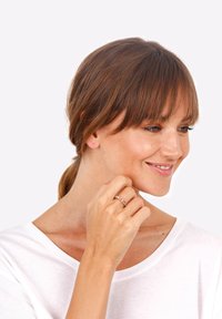 Inel de aur cu design circular legat purtat pe mâna dreaptă. Model cu păr castaniu deschis într-un stil lejer, purtând o cămașă albă, zâmbind.