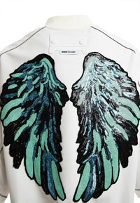 Chaqueta blanca con grandes alas de ángel de lentejuelas en negro y verde azulado en la parte posterior. Textura suave con cuello acanalado y etiqueta de marca visible.