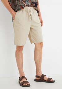 Pantalones cortos beige con cordón, hechos de tejido ligero, que presentan un corte relajado, bolsillos laterales y una textura suave; combinados con sandalias marrones.