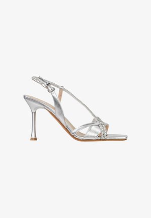 Sandalo argentato con tacco alto, sottili cinturini, fibbia alla caviglia, decorazione con strass e tacco a stiletto sottile su sfondo bianco.