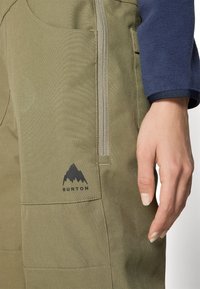 Gröna Burton-byxor med en slät textur, med en sidodragkedja och en svart bergslogo. Närbild på tyget och handen.