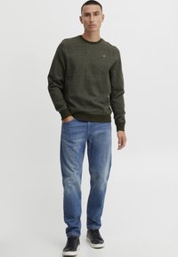 Olijfgroene trui van textiel met een ronde hals en geribbelde manchetten; gecombineerd met lichtblauwe spijkerbroeken en donkere sneakers.