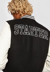 Personne aux cheveux blonds bouclés portant une veste universitaire noire et blanche avec "STARTER" brodé en grandes lettres blanches au dos.