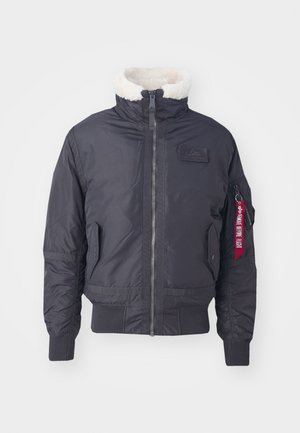 Giubbotto bomber nero isolato con colletto in pelliccia sintetica, zip frontale, tasche laterali, polsini a coste e un'etichetta di stoffa rossa sulla manica.