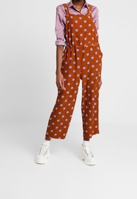 Czarne ogrodniczki w brązowe kropki z jasnopurpurowymi plamkami, szerokie nogawki i regulowane ramiączka. Noszone na fioletowej koszulce, w połączeniu z białymi trampkami.