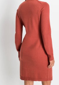 Vestito di maglia color ruggine con maniche lunghe, forma aderente, polsini e orlo a coste, caratterizzato da una superficie liscia e un design minimalista.