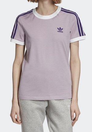 Žena má na sobě světle fialové tričko Adidas s bílým límečkem a manžetami, se třemi tmavě fialovými pruhy na rukávech, doplněné o šedé tepláky.