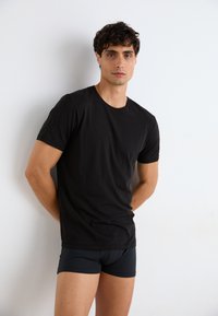 T-shirt en coton noir à manches courtes et encolure ronde, style ajusté. Associé à un boxer noir, modèle en position debout contre un fond blanc.