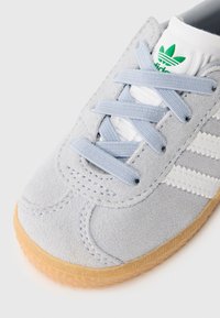 Svetlo sivé semišové tenisky s tromi bielymi pruhmi, modrými šnúrkami a gumenou podrážkou. Majú biely jazyk s detailom loga v zelenej farbe.