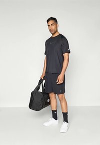 Camiseta y pantalones cortos deportivos negros con logo, usados con calcetines negros y zapatillas blancas. En una mano sostiene una bolsa de viaje negra.