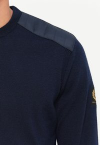 Suéter azul marino con cuerpo de punto texturizado y tela acolchada suave en los hombros; cuenta con un parche de logo en la manga.