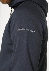 Gros plan d'une personne portant une veste noire avec capuche et le texte brodé blanc "Peak Performance" sur la partie supérieure de la manche.