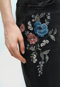 Χέρι που ακουμπάει δίπλα σε μαύρο τζιν από denim με κεντήματα σε μπλε, ροζ και λευκά ανθικά σχέδια κοντά στην τσέπη.