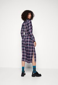 Abito a maniche lunghe a scacchi nei colori blu navy, rosso e bianco. Presenta una chiusura con bottoni sul retro e spacchi laterali. Abbinato a scarpe nere e calze blu con motivo.