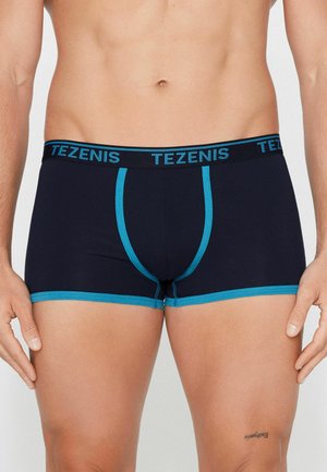 Männlicher Torso trägt marineblaue Boxershorts mit türkisfarbener Einfassung und "TEZENIS"-Logo am Bund, steht mit entspannten Armen.