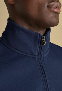 Tums zilas jaku ar rāvējslēdzēju, izgatavota no gludas auduma, ar augstu apkakli un bronzas rāvējslēdzēja akcentu. Tuvplāns pie kakla zonas.