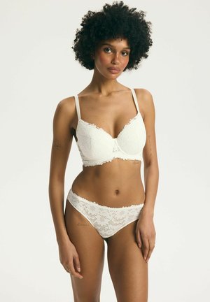 Žena s kučeravými vlasmi, ktorá má na sebe biele čipkované podprsenku a zodpovedajúce nohavičky, stojaca pred svetlým pozadím, otočená čelom s uvoľneným výrazom.