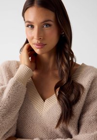 V-hals trui in licht beige, gemaakt van zachte, pluizige stof met geribbelde manchetten en halslijn. Model heeft lang, golvend haar.