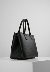 Czarna skórzana torba typu tote o wyraźnym kształcie, z dwoma górnymi uchwytami i subtelną fakturą. Posiada płaskie dno i ochronne metalowe nóżki.
